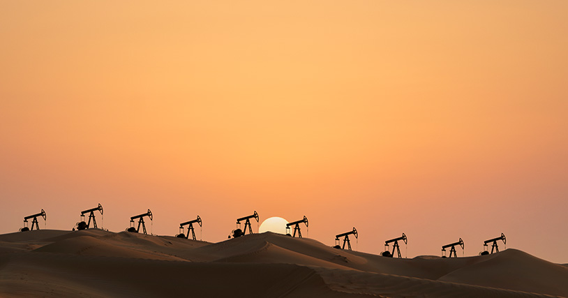A row of oil pumps are silhouetted against a desert sun.