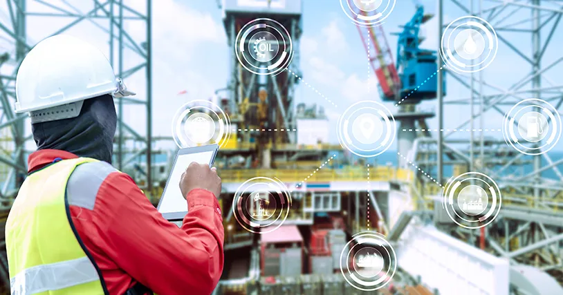 A worker in a hard hat and safety vest on an oil rig holds a tablet, interacting with a digital overlay. 