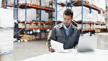 A man in a warehouse looks at documents and a laptop.