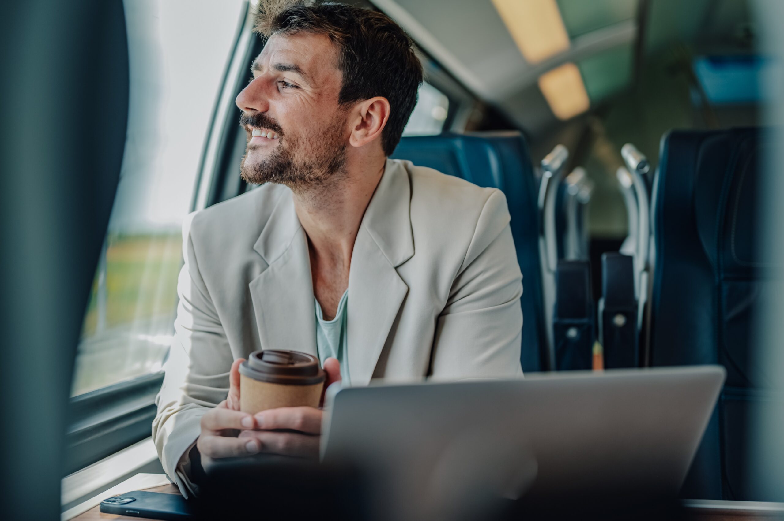A man with a cup of coffee and his laptop looking out the train window