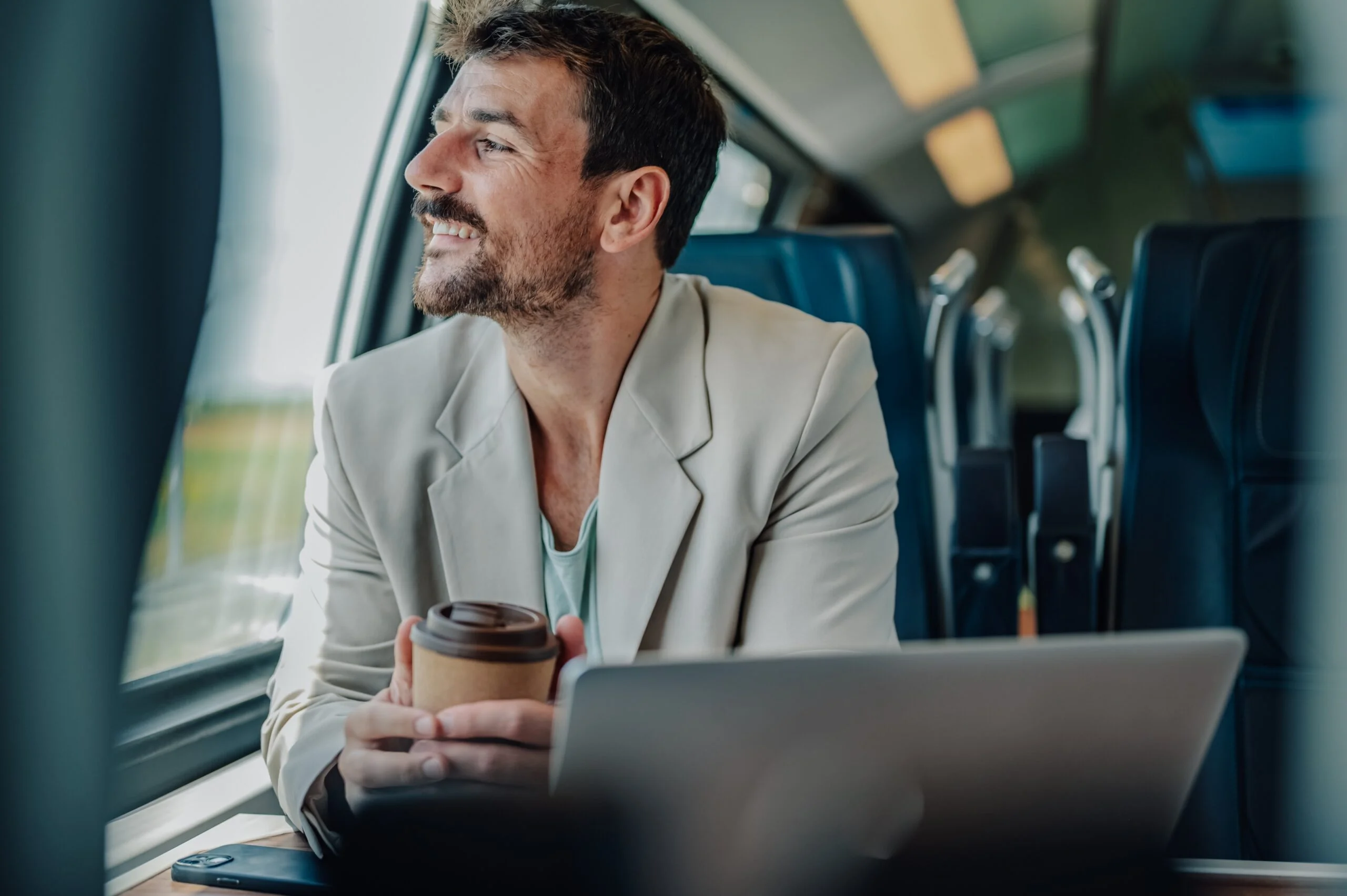 A man with a cup of coffee and his laptop looking out the train window